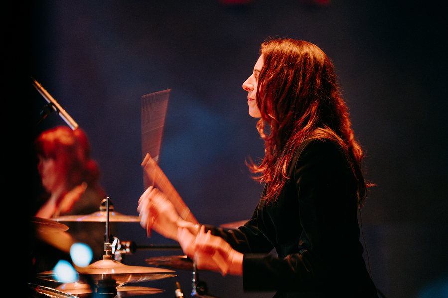 Gail James playing drums, profile, mid-performance, warm stage light against deep blue